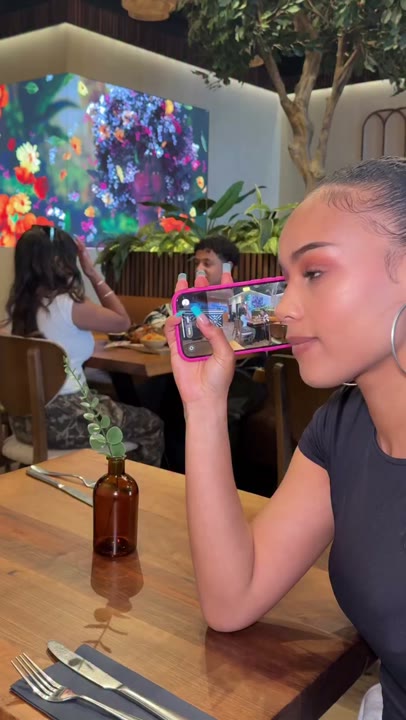 Frame at 0 seconds: Profile framing shows the creator holding a phone beside their face with other diners visible on its screen.