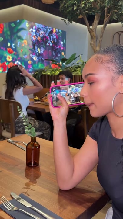 Frame at 1 seconds: Profile framing shows the creator holding a phone beside their face with other diners visible on its screen.