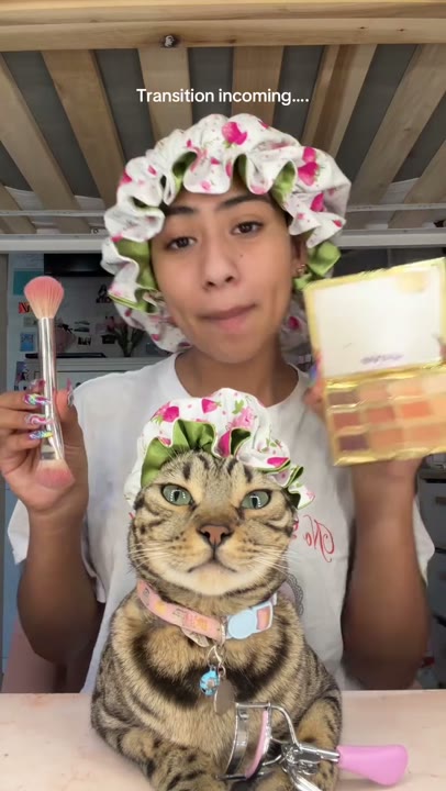 Frame at 2 seconds: The creator raises a makeup brush and open palette beside the cat while lip-syncing.
