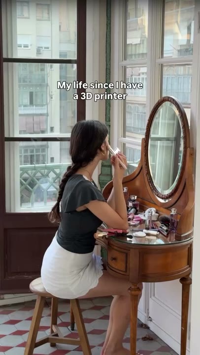Frame at 0 seconds: Makeup routine at a cluttered vanity beneath the 3D-printer ownership premise; the creator pauses and smiles.