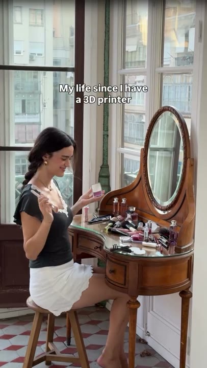 Frame at 2 seconds: Makeup routine at a cluttered vanity beneath the 3D-printer ownership premise; the creator pauses and smiles.